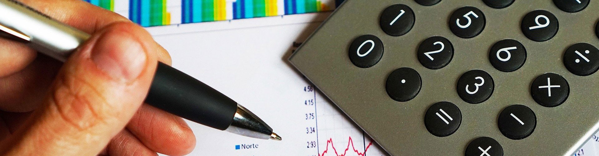 closeup of hand holding pen with calculator and graphs in background