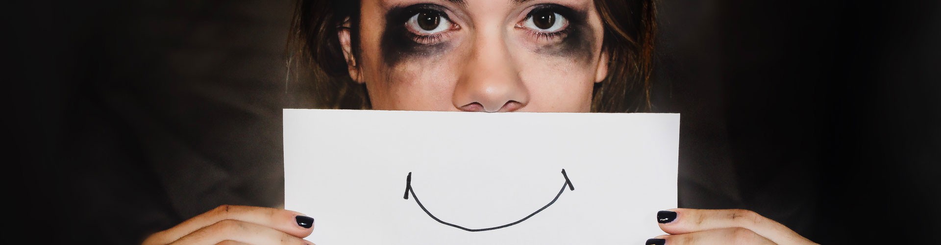 woman with tear stained makeup holds paper with a smile in front of mouth