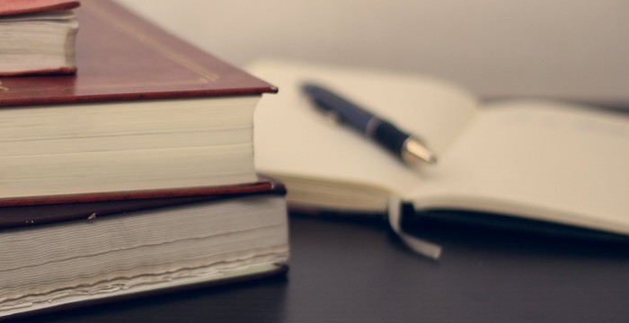A pile of books with a pen resting on an open notebook in the background